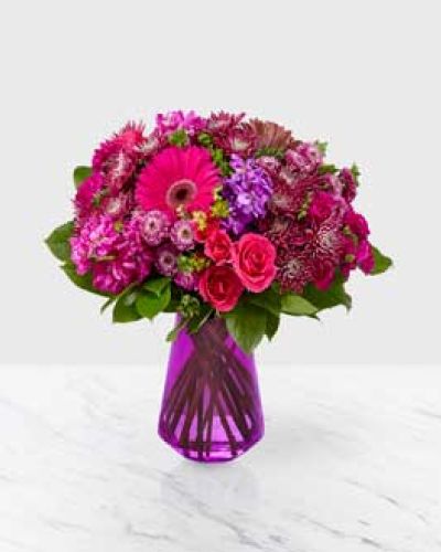Our Blushing Bouquet is sure to turn heads with its explosion of vibrant purple and hot pink tones. There&rsquo;s no comparison to the stunning array of gerberas, mini carnations, spray roses, button poms, and stock, complemented by touches of salal, placed in a vibrant purple glass vase.